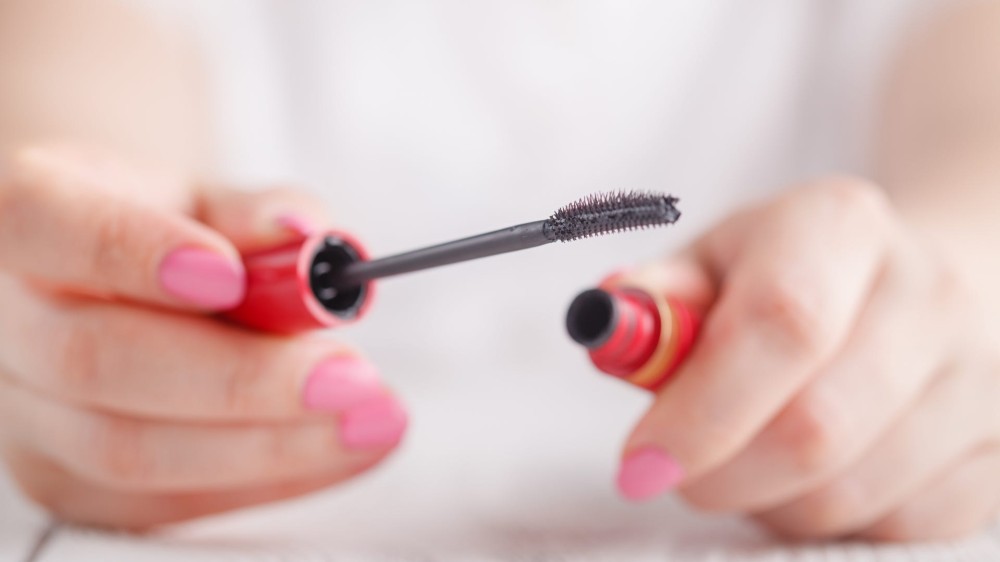 Hands hold a tube of mascara and its applicator wand.