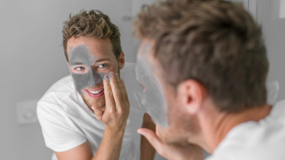 A man applies a charcoal mask to his face while looking in the mirror.