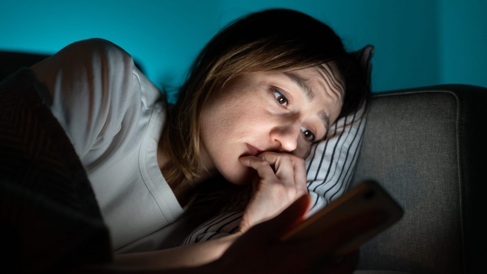 Young anxious woman lying in bed staring at smartphone screen