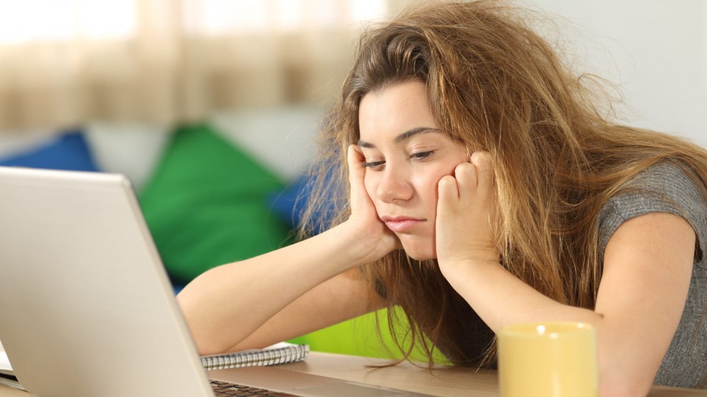 Sleepy student with tousled hair trying to read on line content in a laptop